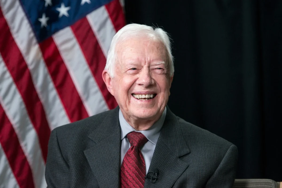 In a press release on his 100th birthday, Jimmy Carter smiles in front of an American flag. Celebrating a century of leadership and accomplishments. 