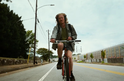 Riding his bike, Teddy (Jesse Plemons) listens to a podcast while on the way to work. Plemons, alongside Aidan Delbis, portrayed conspiracy-obsessed cousins in Yorgos Lanthimos’ “Bugonia.” (photo from Vulture)